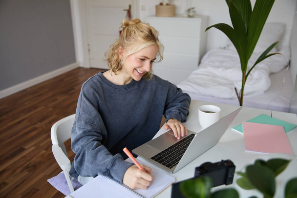 Retrato de una joven sonriente, estudiante, haciendo un curso de aprendizaje a distancia, usando una computadora portátil, estudiando en casa, escribiendo, tomando notas. - Foto, Imagen