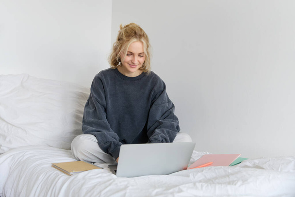 Изображение счастливой молодой женщины, студента электронного обучения из дома, подключиться к онлайн-курс на ее ноутбуке, сидит на кровати с ноутбуками, улыбается и выглядит счастливым. - Фото, изображение