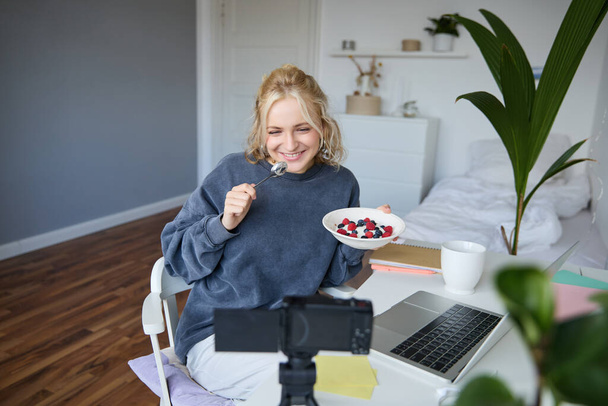 Ritratto di giovane donna sorridente, candida, creatrice di contenuti, mangiare ciotola di dessert e guardare la fotocamera digitale, registrare vlog per seguaci sui social media. - Foto, immagini