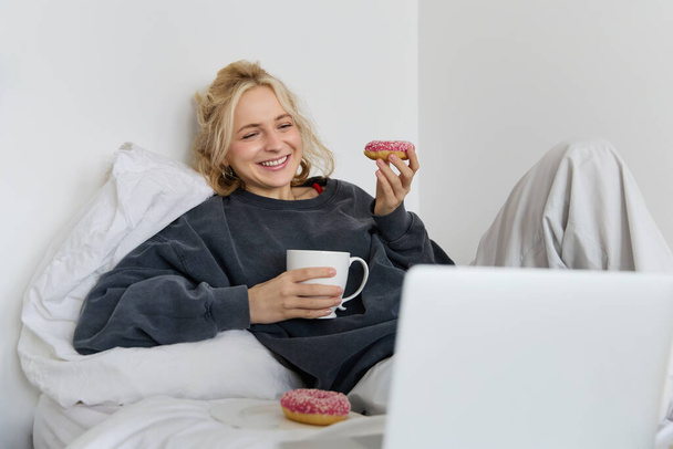 Styl życia i koncepcja ludzi. Szczęśliwa blondynka leżąca w łóżku z jedzeniem, piciem herbaty i jedzeniem pączków, odpoczywająca w domu, spędzająca czas sama, oglądająca program telewizyjny. - Zdjęcie, obraz