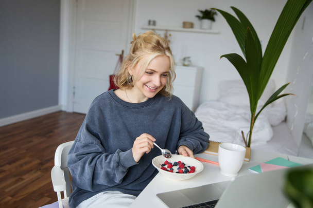 Портрет улыбающейся блондинки, которая ест перед ноутбуком, смотрит видео онлайн за завтраком, наслаждается десертом, сидит в спальне. - Фото, изображение