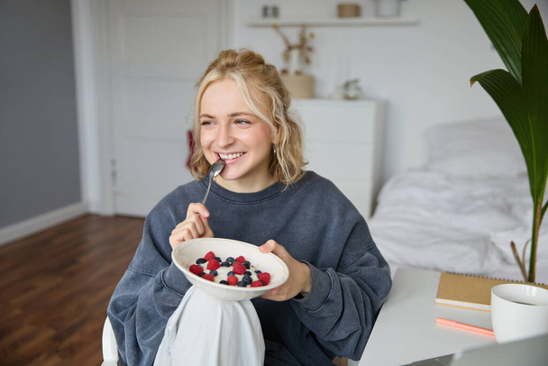Крупным планом портрет улыбающейся, милой блондинки, обедающей в своей комнате, держащей миску с ягодами, кусающей ложку, выглядящей счастливой. - Фото, изображение