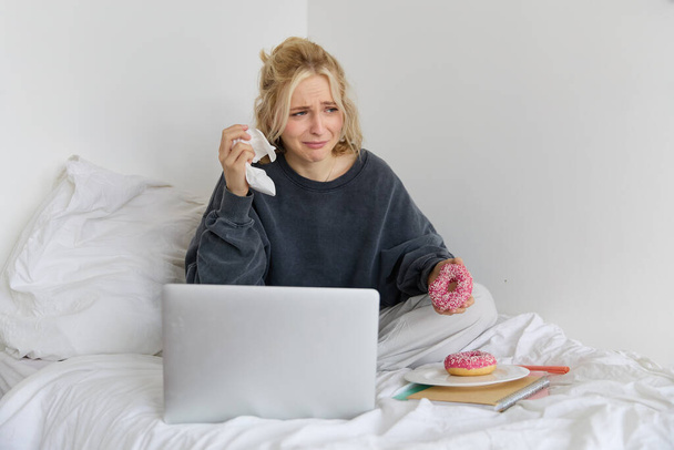 Porträt einer Frau, die auf einem Bett mit Laptop sitzt, Donut isst und aus einer traurigen Filmszene weint. - Foto, Bild