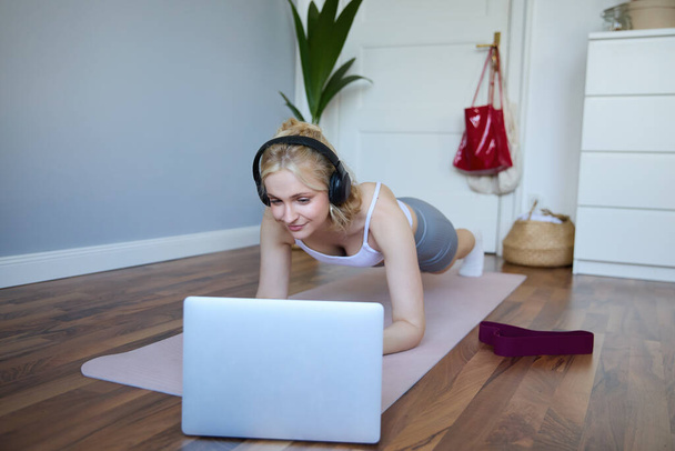 Portrait of young sporty woman doing workout, looking at fitness video on laptop in wireless headphones, standing in plank on yoga mat, following exercise tutorial. - Foto, imagen