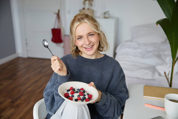 Portrait of smiling blond woman, eating breakfast, holding bowl and spoon, sitting in bedroom, looking happy at camera. Lifestyle concept - Photo, Image