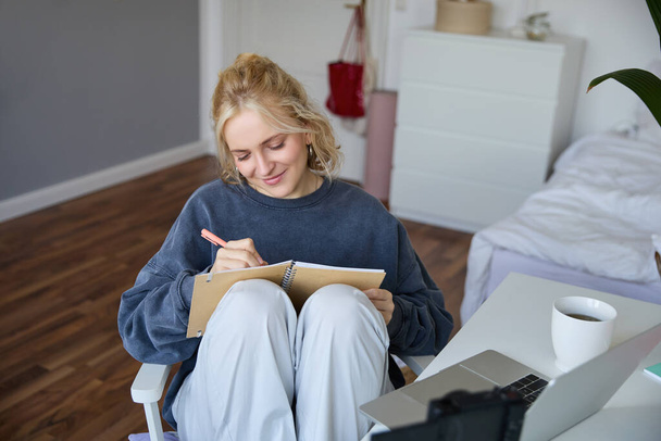 Портрет молодой женщины, пишущей в дневнике, делающей заметки в блокноте, сидящей на стуле перед ноутбуком, занимающейся домашней работой. - Фото, изображение