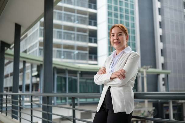 Портрет счастливой азиатской предпринимательницы со скрещенными руками и смотрящей на камеру, стоящую рядом с ее офисом, с большим строительным фоном. Качественное фото. - Фото, изображение