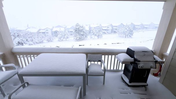 Πυκνά στρώματα χιόνι κουβέρτα μπαλκόνι με έπιπλα εξωτερικού χώρου, προσφέροντας μια γαλήνια θέα ενός προαστιακού τοπίου που περιβάλλεται το χειμώνα αγκαλιάσει. - Φωτογραφία, εικόνα