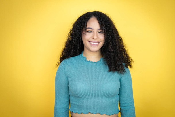 Sarı arka planda sıradan bir kazak giyen Afro-Amerikalı bir kadın mutlu bir yüzü olan ve kendine güvenen bir gülümsemeyle dişlerini gösteren. - Fotoğraf, Görsel