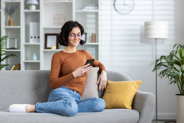 Casual young woman enjoying a leisurely day, smiling as she uses her smartphone on a comfortable couch in a well-lit living room. - 写真・画像