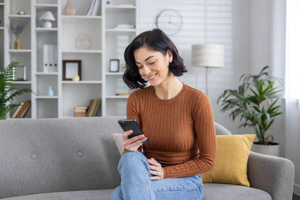 A cheerful young woman engaged with her smartphone, comfortably seated on a modern sofa in a well-lit living room. - Foto, afbeelding