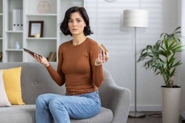 A woman holds a smartphone and credit card feeling confused and concerned about an error during an online ,transaction while sitting on the couch at home. - Φωτογραφία, εικόνα
