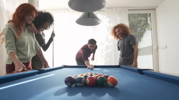 Молодий чоловік в повсякденному одязі розбиває стійкі басейни на більярдний стіл, оточений щасливими різноманітними друзями в ігровій кімнаті під час вихідного дня вдома - Кадри, відео