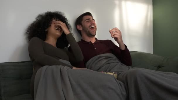 Laughing diverse young couple on cozy sofa eating popcorn while enjoying comedy movie in living room at home during weekend day - Footage, Video