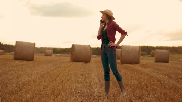 Kobieta pracująca na farmie w gumowych butach na polu pszenicy, agrarna kobieta rozmawiająca przez telefon komórkowy o plonach pszenicy na tle okrągłych bel. Praca w rolnictwie rolnym i produkcja ekologiczna - Materiał filmowy, wideo
