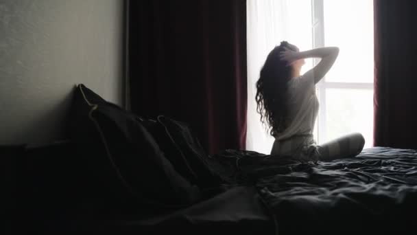 Giovane donna seduta sul letto, guardando fuori dalla finestra mentre si tocca i capelli ricci a casa. Al rallentatore. Stile di vita concetto - Filmati, video