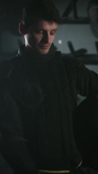 Siyah üniformalı genç silahşörün dikey portresi karanlık odada durup kameraya bakarken omuz omuza yükseliyor. - Video, Çekim