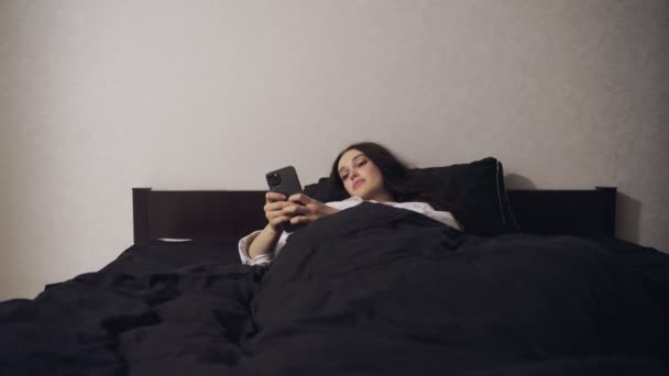 Молода жінка використовує смартфон і спілкується перед сном в спальні вдома. Повільний рух. Концепція стилю - Кадри, відео