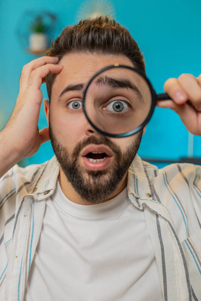 Caucasian young freelancer man holding magnifying glass near face, looking into camera with big zoomed funny big eye, searching, analyzing. Bearded guy sitting at home office table workplace. Vertical - Photo, Image