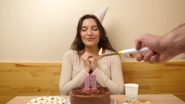 The girl is sitting in front of a table with a festive cake, in which a candle in the form of the number 17 is burning, which she blows out. Birthday celebration concept. - Footage, Video
