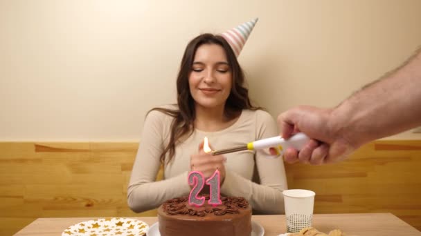 The girl is sitting in front of a table with a festive cake, in which a candle in the form of the number 21 is burning, which she blows out. Birthday celebration concept. - Footage, Video