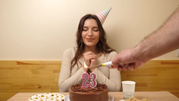 The girl is sitting in front of a table with a festive cake, in which a candle in the form of the number 23 is burning, which she blows out. Birthday celebration concept. - Footage, Video