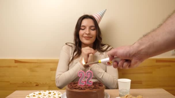 The girl is sitting in front of a table with a festive cake, in which a candle in the form of the number 22 is burning, which she blows out. Birthday celebration concept. - Footage, Video