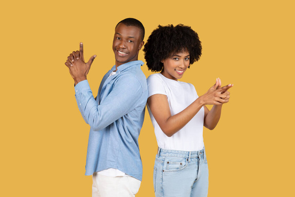 Back-to-back millennial African American couple confidently showing hand signs with playful expressions, dressed in casual attire against a uniform yellow background, studio - Photo, Image