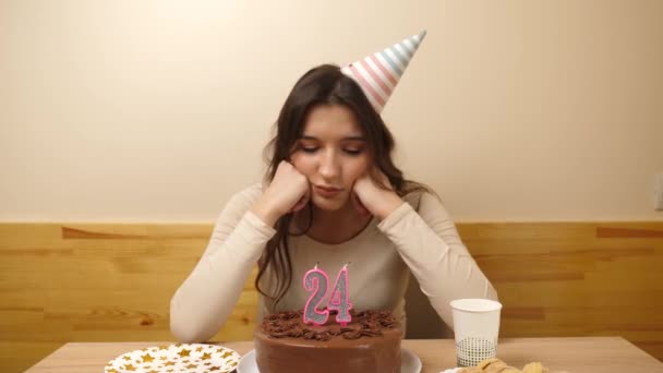 Расстроенная девушка садится перед столом с праздничным тортом, в котором загорается свеча в виде цифры 24. Концепция дня рождения  - Кадры, видео