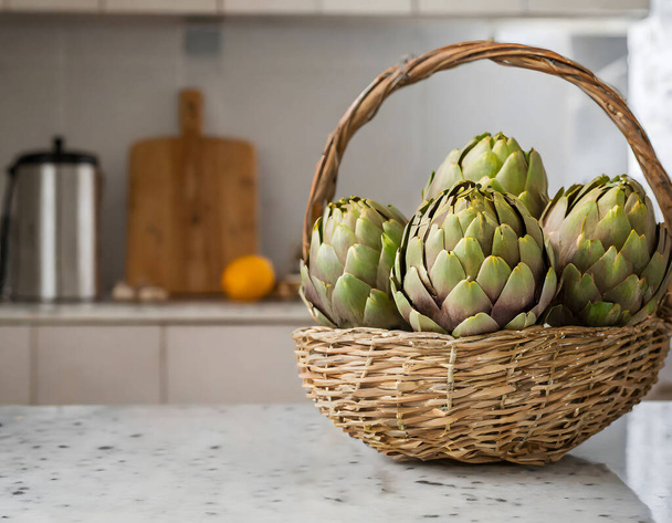 Un ramo de alcachofas verdes en una cesta de mimbre rústica, colocadas en una encimera de cocina con utensilios de cocina en el fondo - Foto, imagen