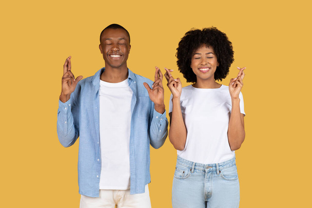 A cheerful young African American couple wearing casual clothing are crossing their fingers with eyes closed against a yellow background, hoping for good fortune, studio. Hope, win - Photo, Image