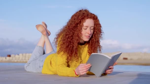 Een roodharige jonge vrouw die buiten een boek leest. Leerling op de grond. - Video