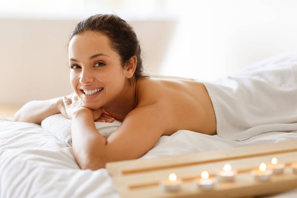 Hymyilevä nainen kylpylässä makaamassa käärittynä valkoiseen pyyhkeeseen, lepäämässä ja odottamassa rentouttavaa hierontaa keskellä palavia kynttilöitä, edistäen hyvinvointia ja kehon nuorentavaa hoitoa lomakohteessa - Valokuva, kuva