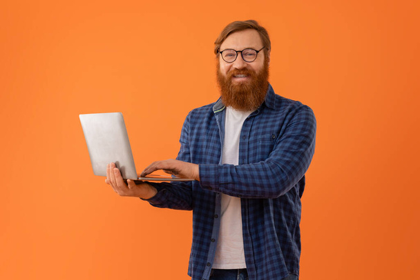 Kırmızı saçlı sakallı iş adamı dizüstü bilgisayarda çalışıyor, dijital profesyonelliği ve serbest kariyer fırsatlarını gösteren kameralara gülümsüyor, turuncu arka planda - Fotoğraf, Görsel