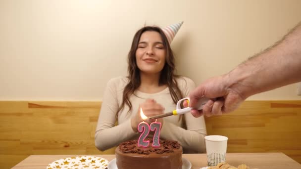 The girl is sitting in front of a table with a festive cake, in which a candle in the form of the number 27 is burning, which she blows out. Birthday celebration concept. - Footage, Video