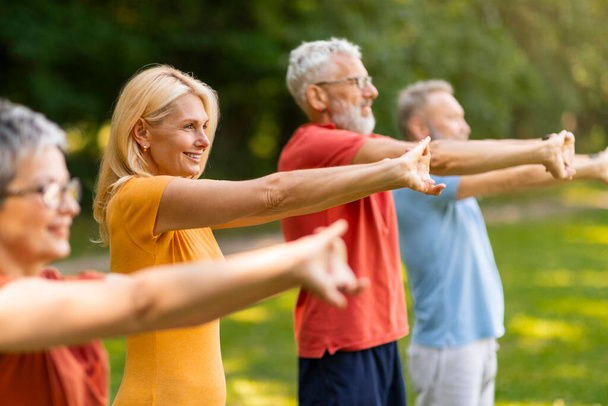 Grupa szczęśliwych seniorów wykonująca ćwiczenia rozciągające podczas treningu na świeżym powietrzu, uśmiechnięci starsi mężczyźni i kobiety wyciągający ramiona do przodu, ćwiczący razem jogę, cieszący się aktywnym stylem życia na emeryturze - Zdjęcie, obraz