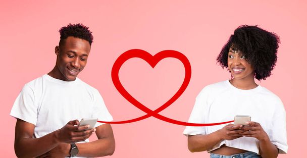 スマートフォンに囲まれた男性と女性は,ソフトピンクの背景に設定された大きな赤いハートを介して接続を共有し,仮想愛情を描きます - 写真・画像