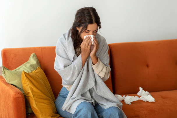 Hasta, genç bir kadın battaniyeye sarılmış, burnunu kağıt mendille siliyor, hasta bir kadın oturma odasında kendini kötü hissediyor, soğuk algınlığı veya grip belirtileri gösteriyor. - Fotoğraf, Görsel