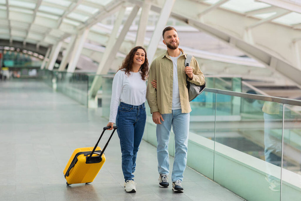 Retrato de pareja joven positiva caminando en el aeropuerto con equipaje, feliz hombre y mujer milenial llevando maleta y mochila pasando por el pasillo en la terminal, disfrutando viajando juntos - Foto, imagen