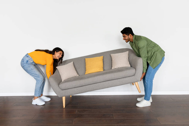 Glückliche indische Ehepartner, die ihr graues Sofa im Zimmer über weißem Wandhintergrund in ihrem neuen Zuhause bewegen, in eine neue Wohnung ziehen, volle Länge, freier Raum - Foto, Bild