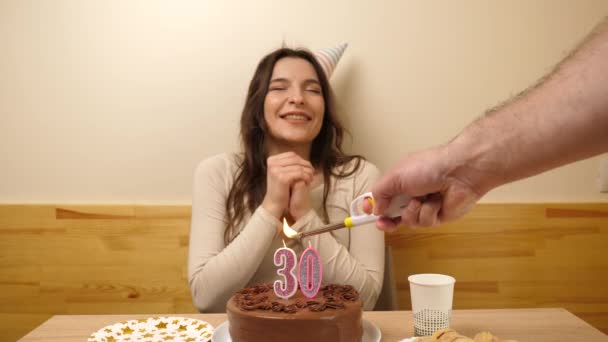 The girl is sitting in front of a table with a festive cake, in which a candle in the form of the number 30 is burning, which she blows out. Birthday celebration concept. - Footage, Video