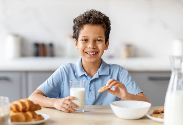 Boldog fekete fiú tejet iszik és sütit eszik a konyhában, poharat tart, mosolyog. Vidám gyerek tartja egészséges táplálkozás, egyre kalcium, vitaminok, probiotikumok a növekedés tejtermékek - Fotó, kép