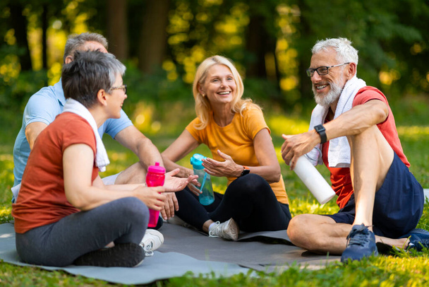Gioioso anziani appassionati di yoga godendo di una chiacchierata amichevole mentre seduti su stuoie all'aperto, anziani felici in possesso di bottiglie d'acqua, uomini e donne anziani rilassarsi dopo una sessione rinfrescante nel parco estivo - Foto, immagini