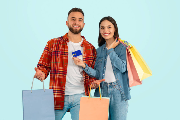 ショッピング。 ハッピーミレニアルカップルは彼らの購入を披露し,デビットカードで支払い,青い背景を持つスタジオに立って,季節の販売とファッション小売トレンドを楽しんで,一緒に微笑んで - 写真・画像