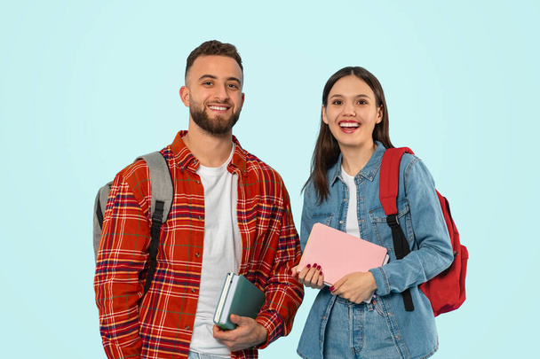 Moderne Studenten. Ein fröhliches junges Paar posiert lässig mit Rucksäcken, Schulbüchern und Gadgets über einer blauen Wand im Studio und lächelt in die Kamera, während es das moderne College-Leben repräsentiert. Bildungskonzept - Foto, Bild
