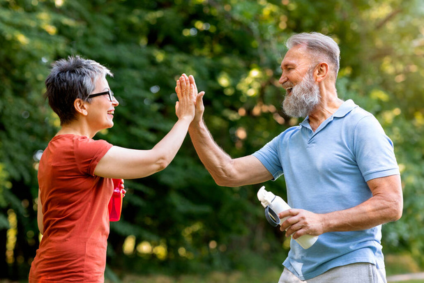 Hombre y mujer mayores energéticos que se dan choca los cinco al aire libre, personas mayores alegres felices celebrando los objetivos de fitness en el soleado parque verde, sosteniendo botellas de agua en la mano, vista lateral - Foto, Imagen