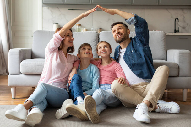 Uśmiechnięta czteroosobowa rodzina siedząca na podłodze, tworząca symboliczny dom z połączonymi dłońmi, szczęśliwi młodzi rodzice i nastoletnie dzieci oznaczające jedność i schronienie w salonie - Zdjęcie, obraz