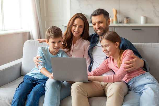 Wesoła młoda rodzina z dziećmi bawiącymi się z laptopem w domu, oglądającymi razem zdjęcia. Szczęśliwi rodzice i ich dwoje nastolatków korzystających z komputera podczas siedzenia na kanapie w salonie, wolna przestrzeń - Zdjęcie, obraz