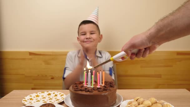 El niño se sienta frente a una mesa con un pastel festivo y velas, las sopla y recibe regalos. Concepto de celebración de cumpleaños. - Metraje, vídeo