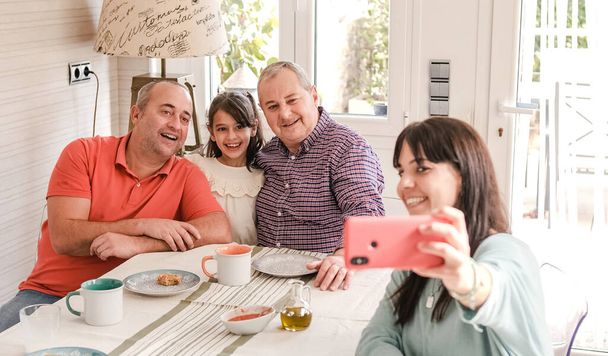 Усміхаючись середнього віку гей-пара з онукою і дочкою, дивлячись на екран мобільного телефону, сидячи за обіднім столом під час сніданку і приймаючи селфі разом вдома - Фото, зображення